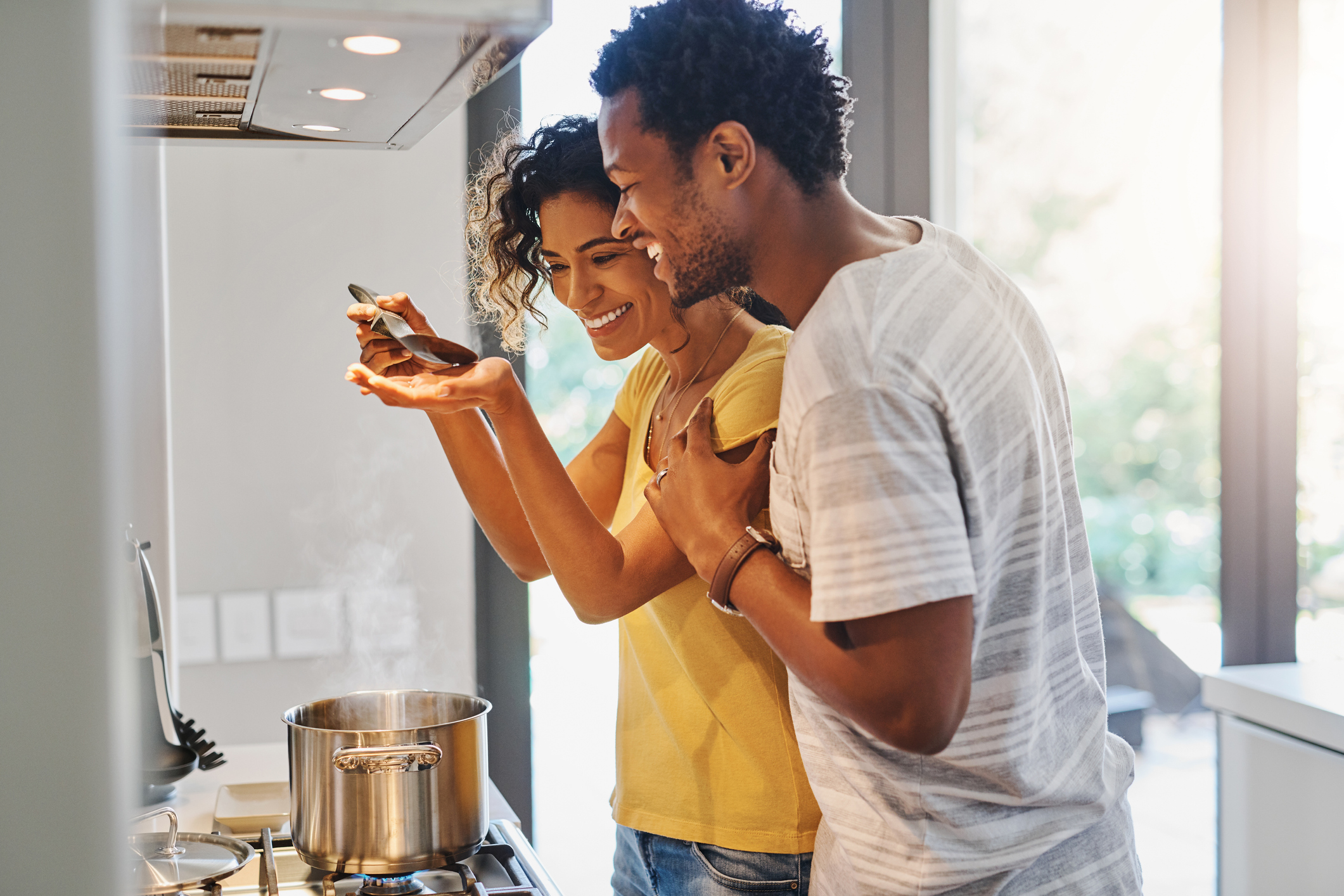 A man and woman smile and cook while trying food from a ladle