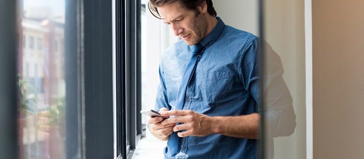 man looks at phone while standing next to window