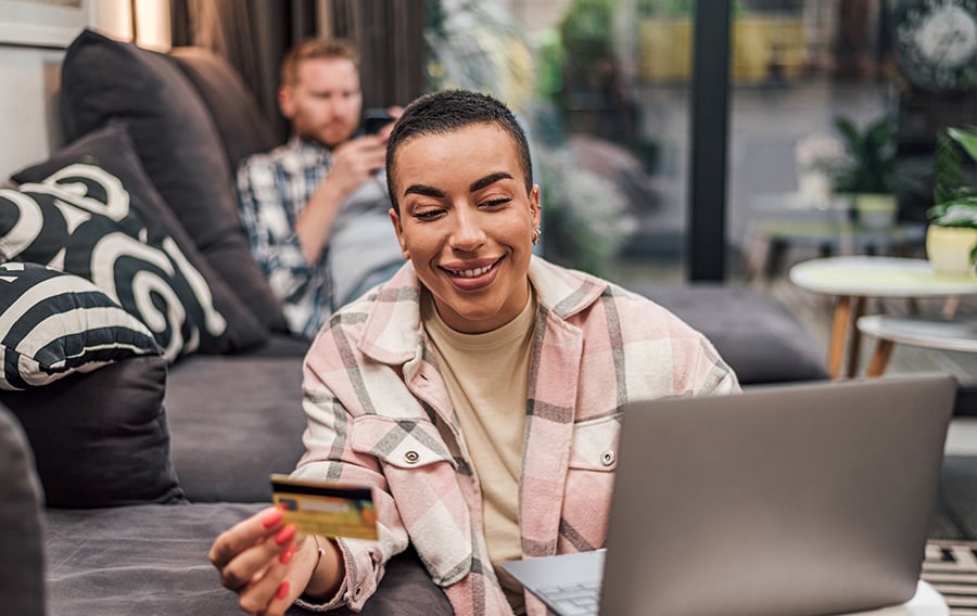 Person sits in a living room and smiles while holding a credit card and looking at a laptop