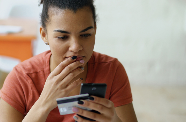 A young woman looks contemplatively at her phone while holding a credit card