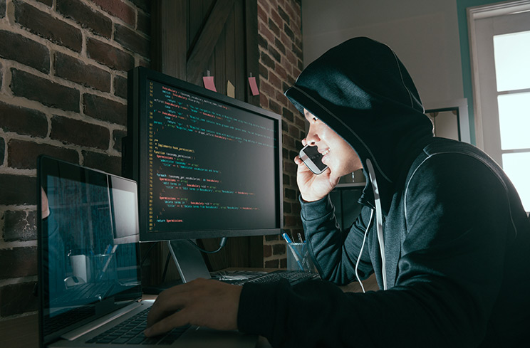 hooded hacker on phone smiling while looking at computer screen
